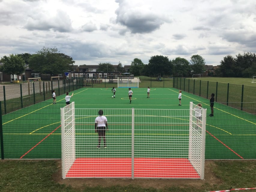 A Multi Use Games Area that has been surfaced with a green polymeric surfacing. 9 Children are playing a match of football and the picture is taken from the goal.