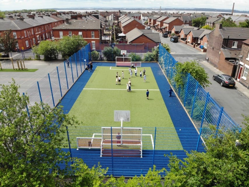 A MUGA pitch that has a wetpour surface going around the border of the pitch. Children are playing a game of football on the pitch.