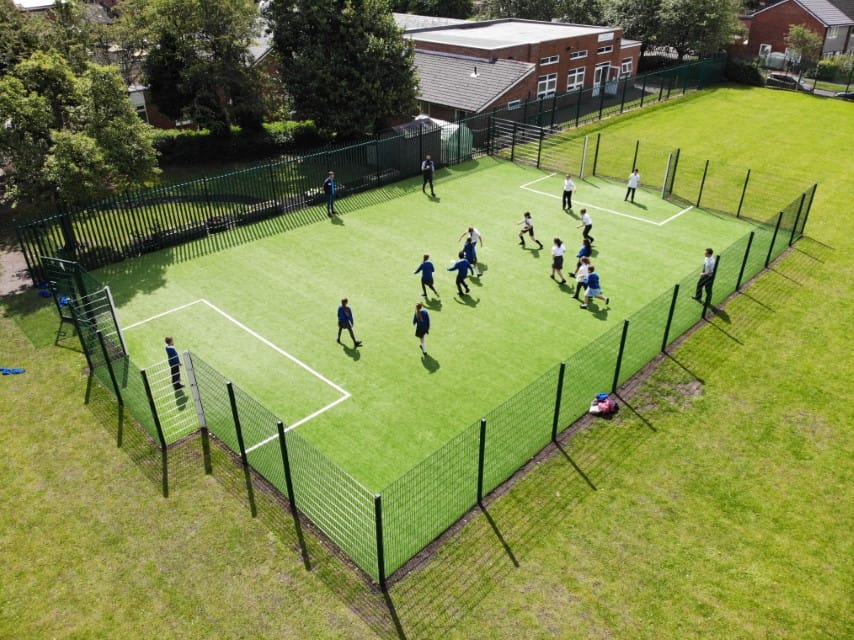 A small Multi Use Games Area which is green with two box markings, showing the goal keepers area. The children are playing football against each other. The MUGA has a green fence going around it.