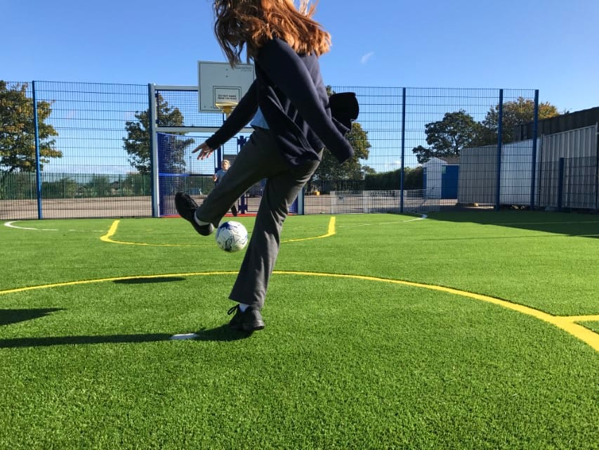 A close up shot of a child kicking a football towards a goal on an artificial grass MUGA Pitch. The photo is taken from the back of the child.