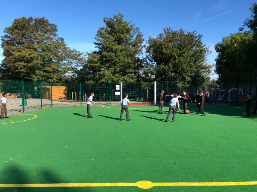 A picture showing the synthetic grass surfacing of a MUGA. A group of children have separated into teams by taking off (or keeping on) jumpers. They are playing football on green synthetic grass with line markings.