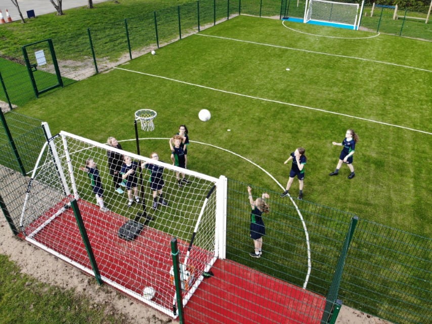 8 young girls and a teacher are playing netball on an artificial grass MUGA Pitch. The girls are dressed in P.E. Uniforms. One child is shooting the netball in to the netball hoop.