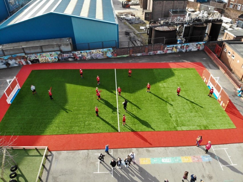 An artificial grass MUGA with a single line marking down the middle. An outline of red wetpour can be seen going around the outside. 12 children are playing football against each other with one teacher being the referee. The photo is from a drone 