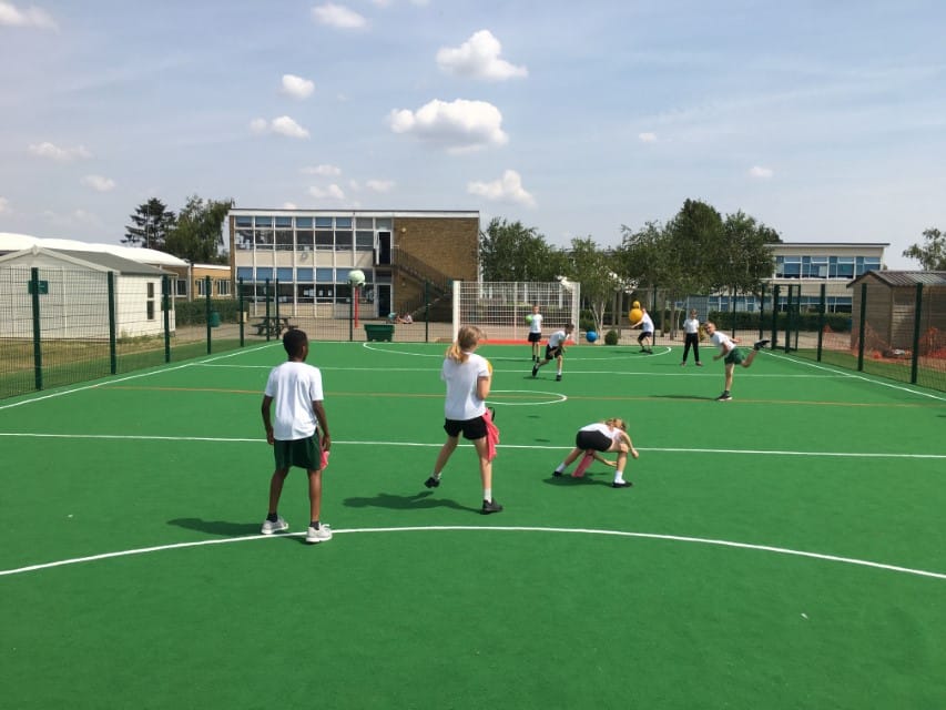 8 Children are playing dodgeball on a MUGA pitch that we installed. The muga is green with white line marking for football on it, The kids are dressed in P.E. kits as they get some exercise through dodgeball.