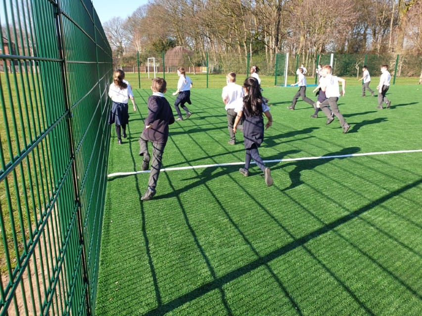 A picture of children running on a MUGA. The kids are wearing school uniform as they run on artificial grass. The MUGA is surrounded with a green fence.