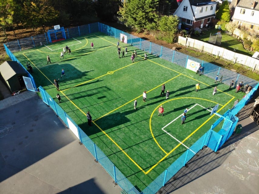MUGA pitch which has blue fencing going around it. Yellow line markings are showing the area for football. A large amount of kids are playing sports on the MUGA.