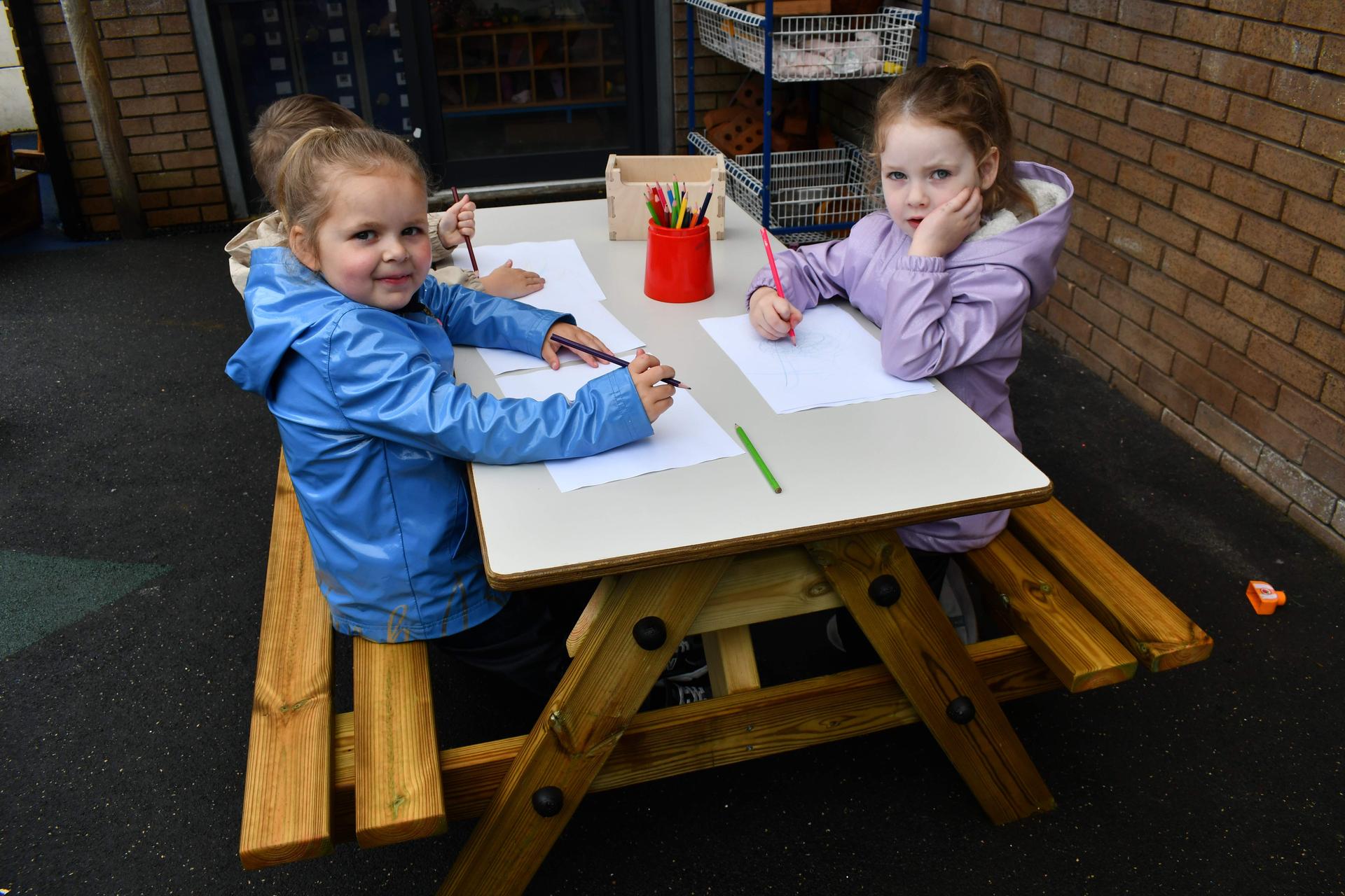 3 children sat on the Early Years Picnic Table. The children are looking at the camera, smiling, as they are drawing on paper which is placed on top of the EYFS Picnic table.