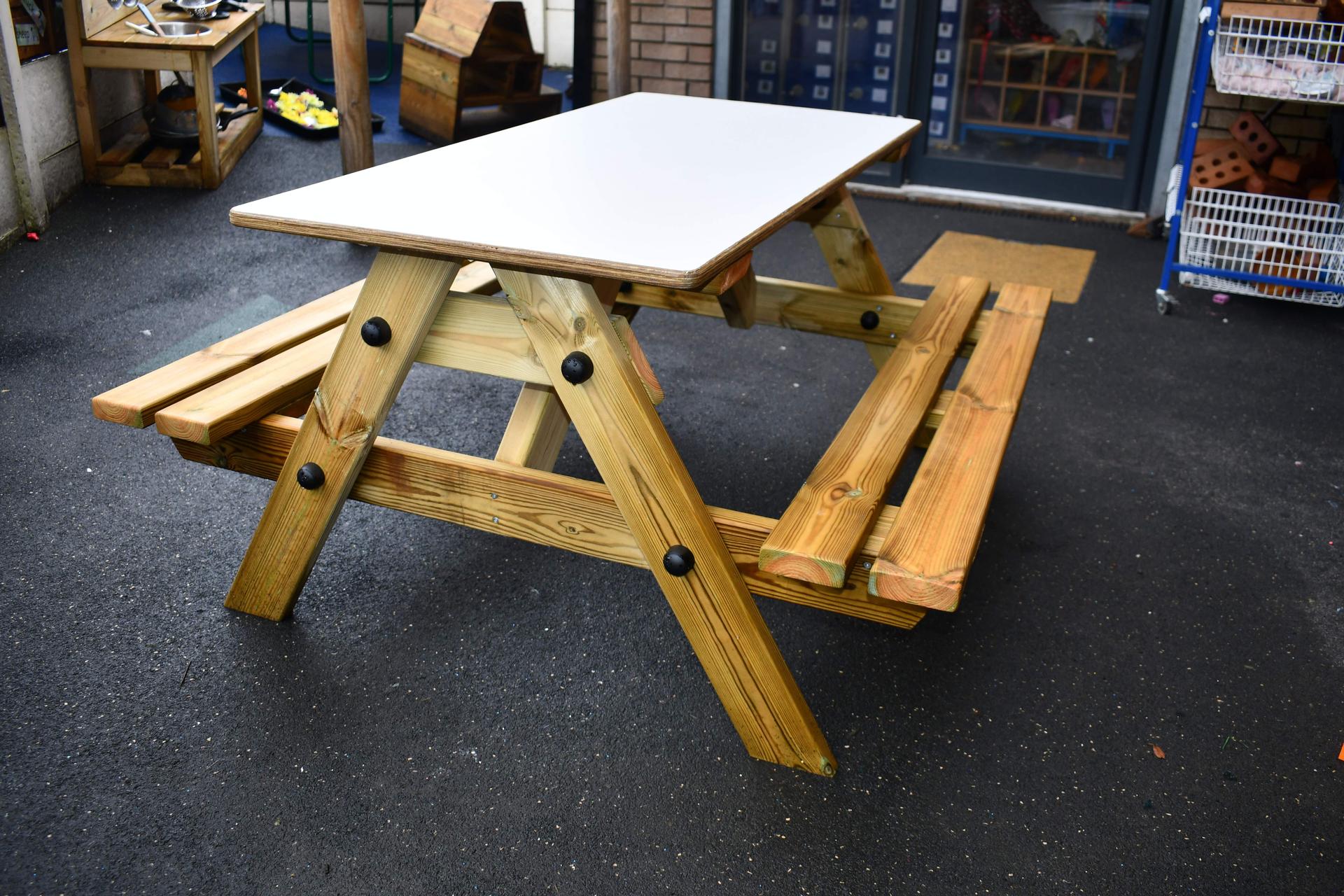 The EYFS Bench Assembled. It is set up on tarmac with a brick background. The top of the bench is grey with the rest being natural wood.