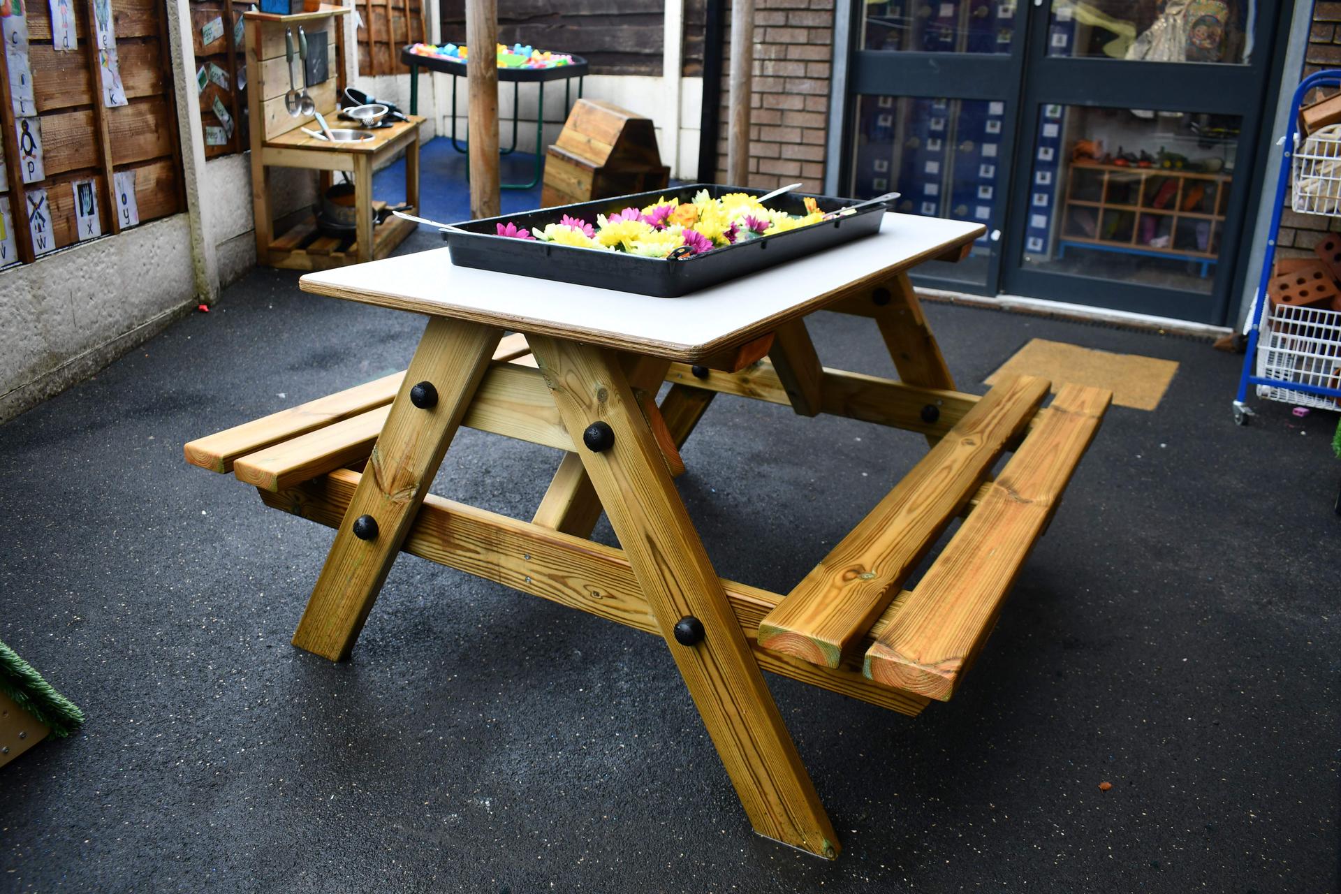 The Early Year Picnic Table is assembled and placed on top of tarmac. A tuff tray full of flowers and green goo is placed on the grey surface of the EYFS table.