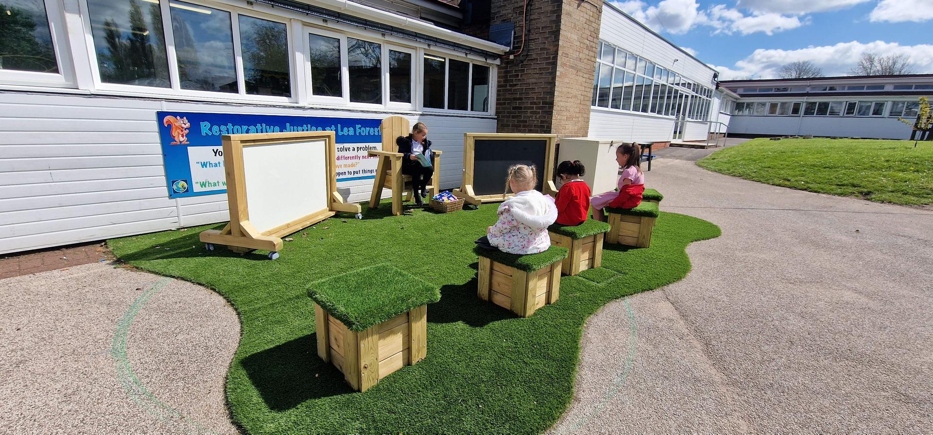 3 children are sat on wooden blocks with artificial grass on top of them. A child is sat on a big wooden chair, with a whiteboard and chalkboard on either side of them.