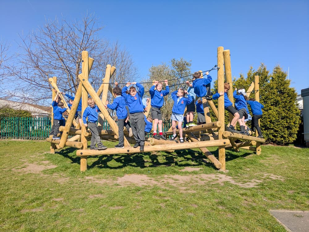 A group of school children in blue jumpers playing on a large wooden climbing frame in a playground.