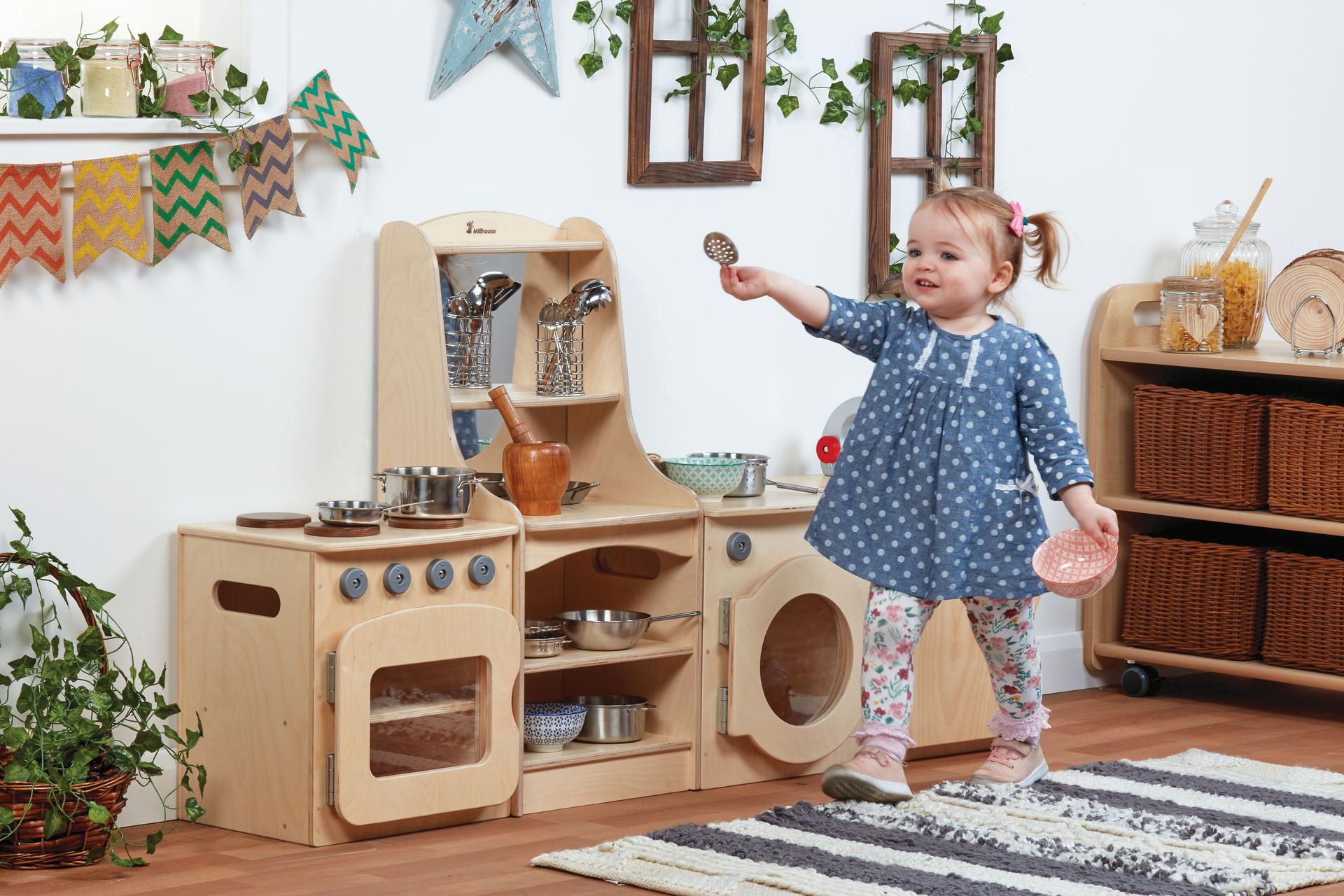 A young girl playing with the Natural Kitchen Set of 4 - Cooker, Sink, Washer, Storage Dresser (H400mm), inside a classroom.