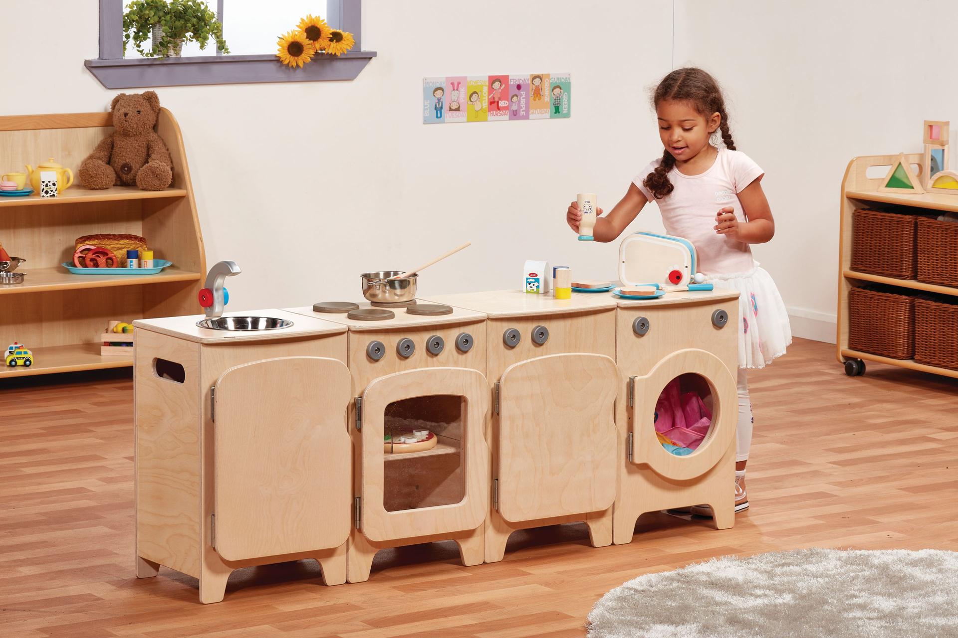 A pre-school aged girl is playing with the Natural Kitchen Set of 4 - Cooker, Sink, Washer, Fridge (H550mm), inside a classroom.
