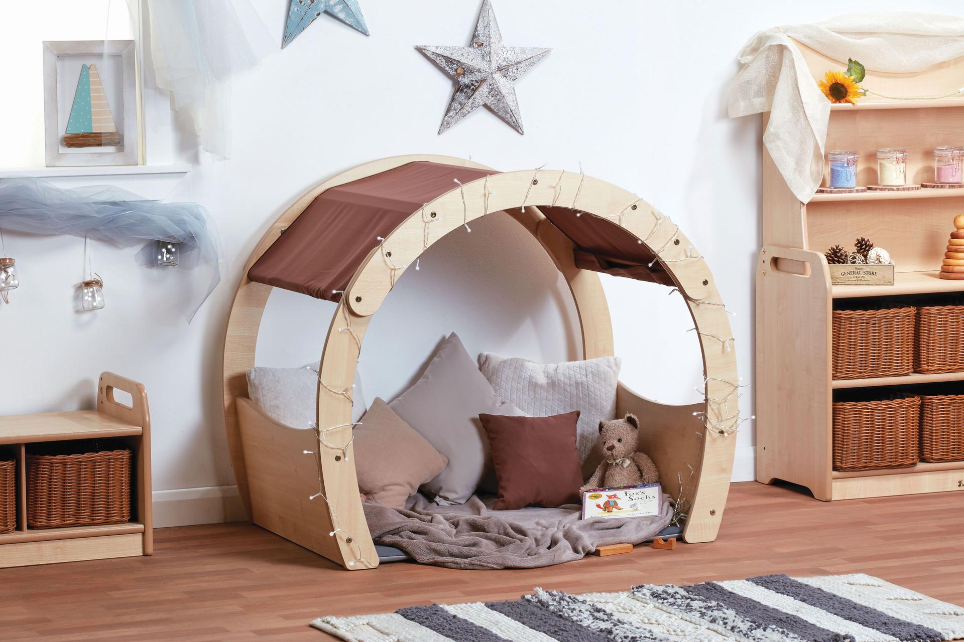 A wooden arch with brown fabric top within a nursery setting, filled with cushions and blankets.