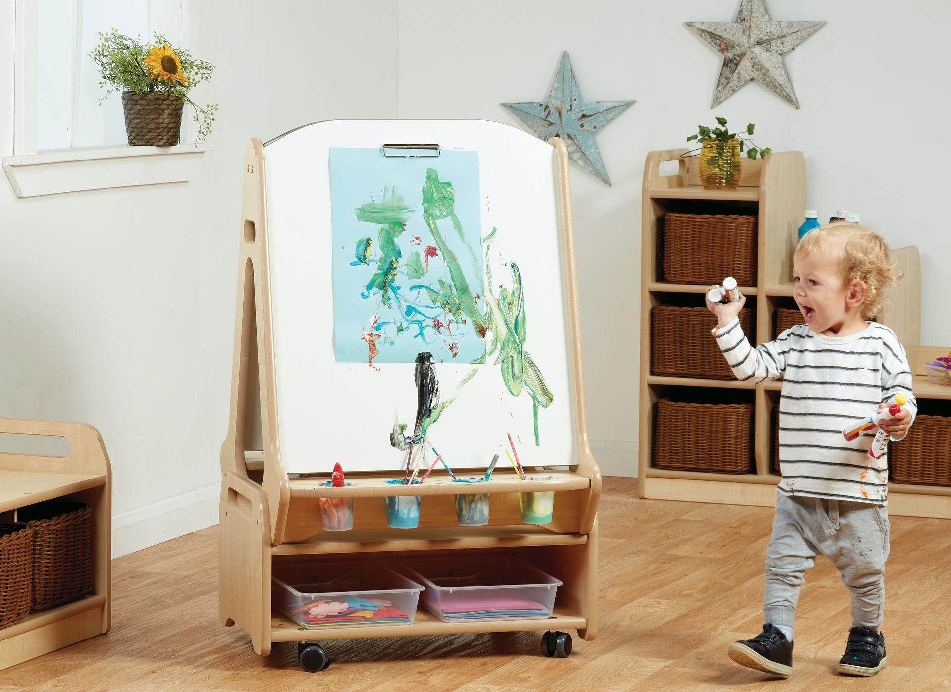 A young boy is painting using the Double-sided 2 Station Whiteboard Easel with Low Storage Trolley.   