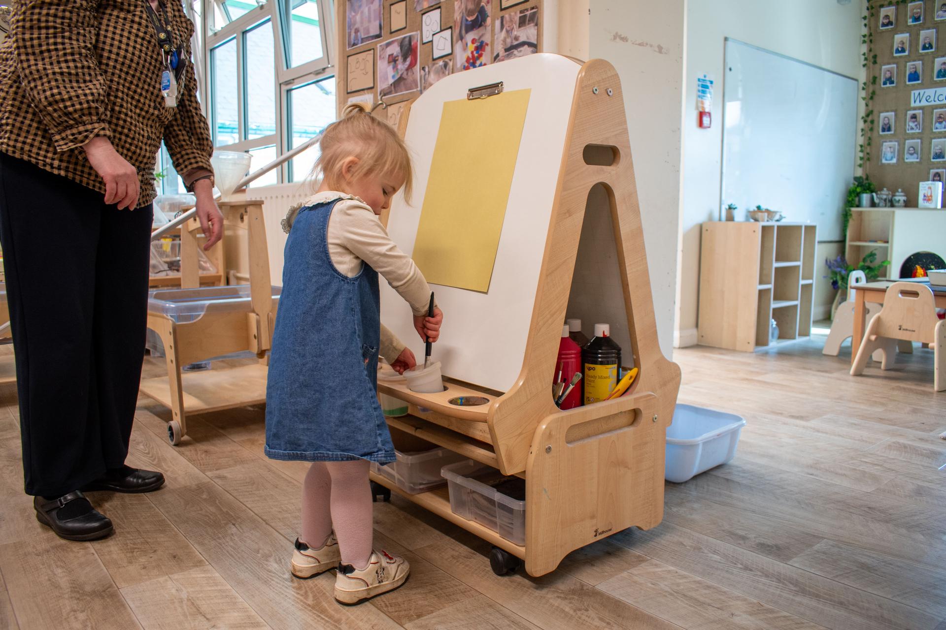 A Nursery practitioner helps a young girl paint on the Millhouse Easel
