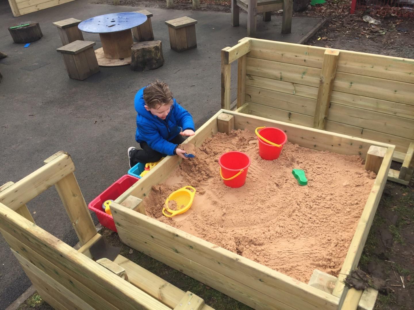 A boy is digging around in the small covered sand box as he uses a plastic shovel and bucket.