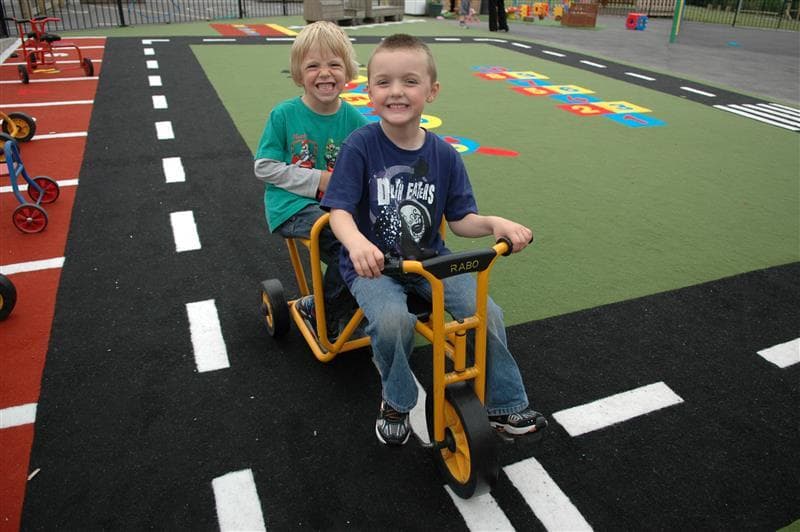 Two young boys riding scooters on the roadway in their playground at school