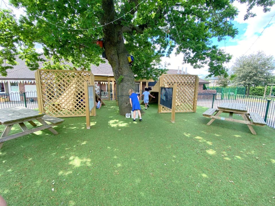 a child plays under a tree on our artificial grass between two trellises and a picnic table- the grass is part of a fenced off area of a school playground