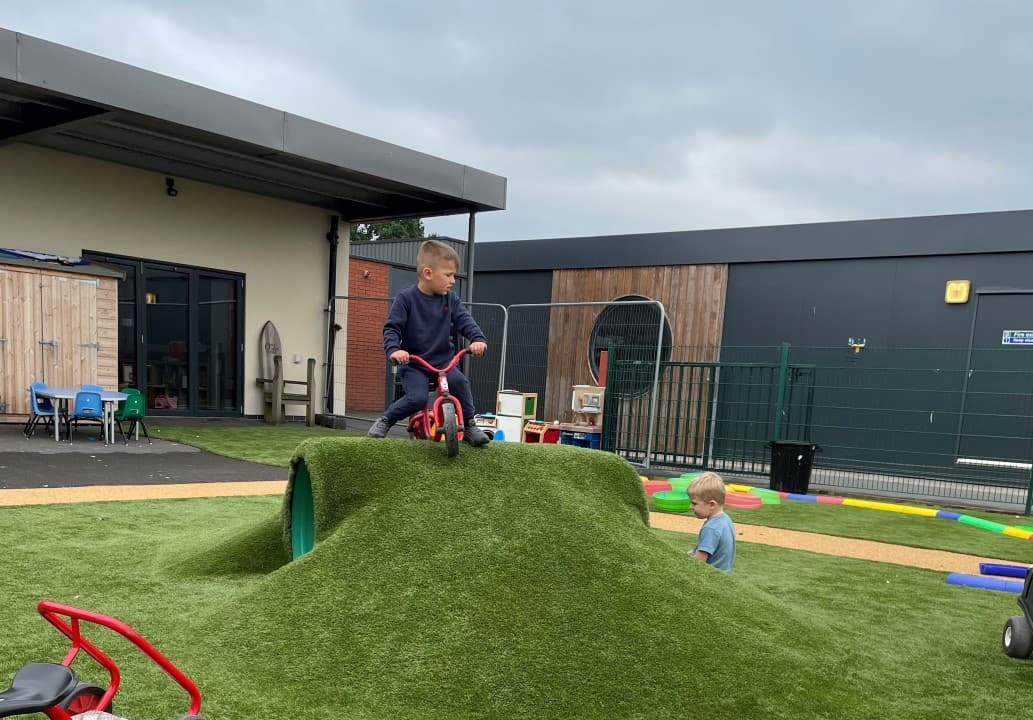 a child in blue school uniform sits on a red bike on top of a green artificial grass playground mound with a tunnel below