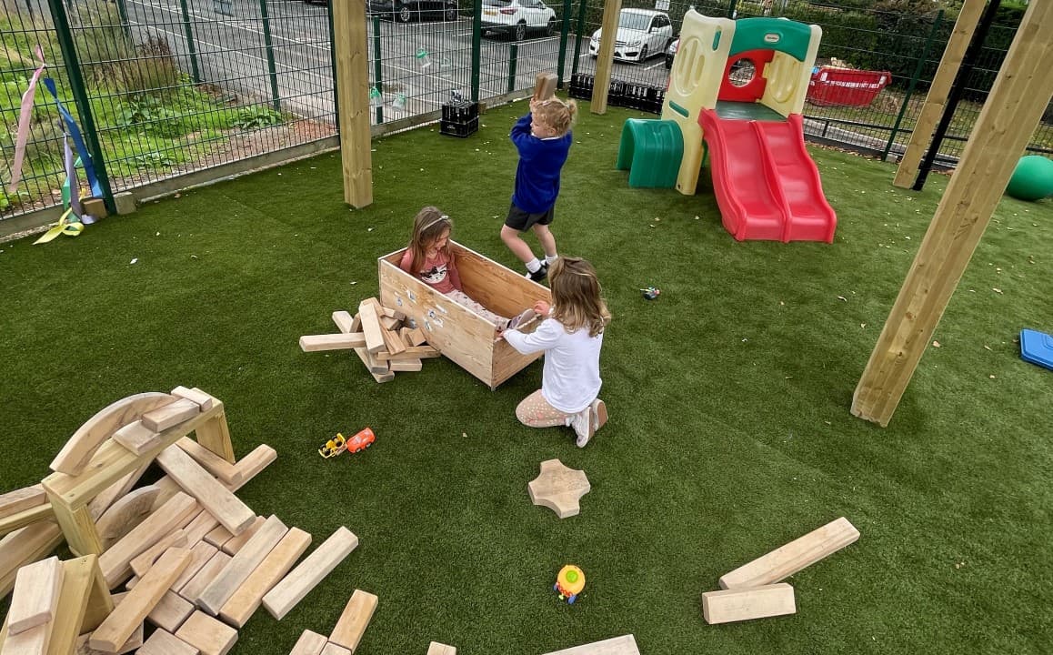 three girls sit and play with wooden blocks on the green artificial grass surfacing