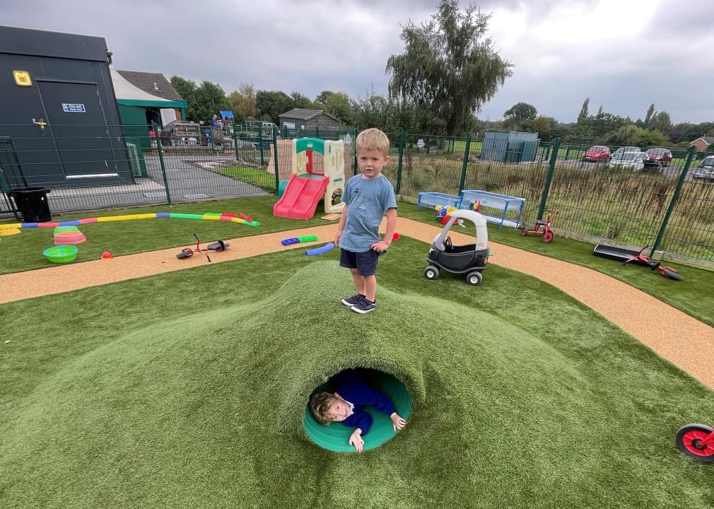 a child in the green artificial grass tunnel with a child stood on top of the mound above