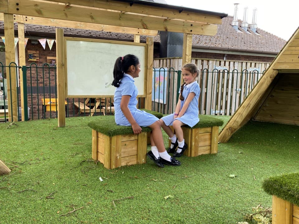 two little girls sat conversing on wooden blocks topped with artificial grass for comfort, there is a giant whiteboard behind them and aritficial grass beneath them