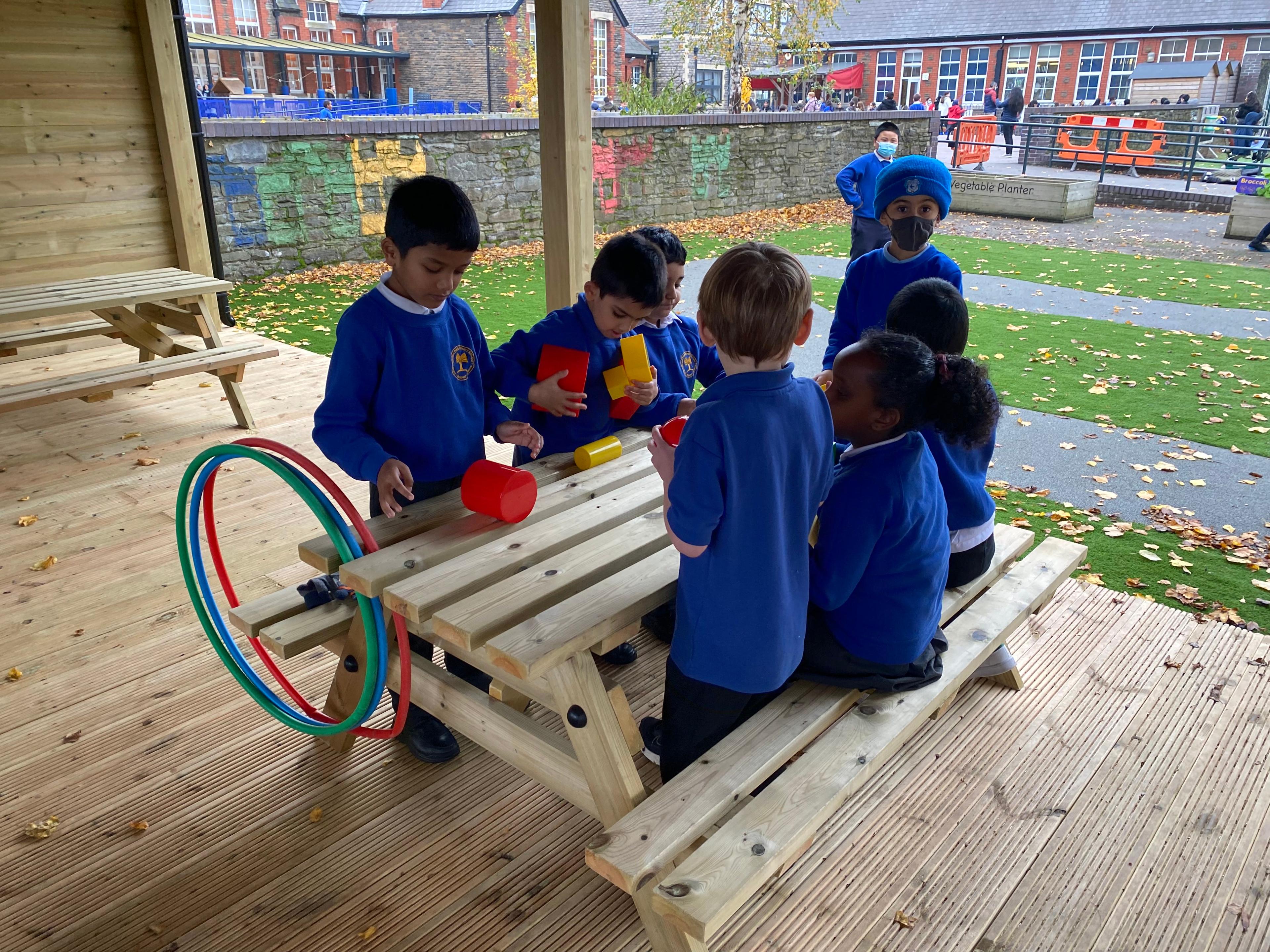 5 children sitting on the picnic bench playing with plastic cups and toys, there are hoops hanging off the side of the bench. 