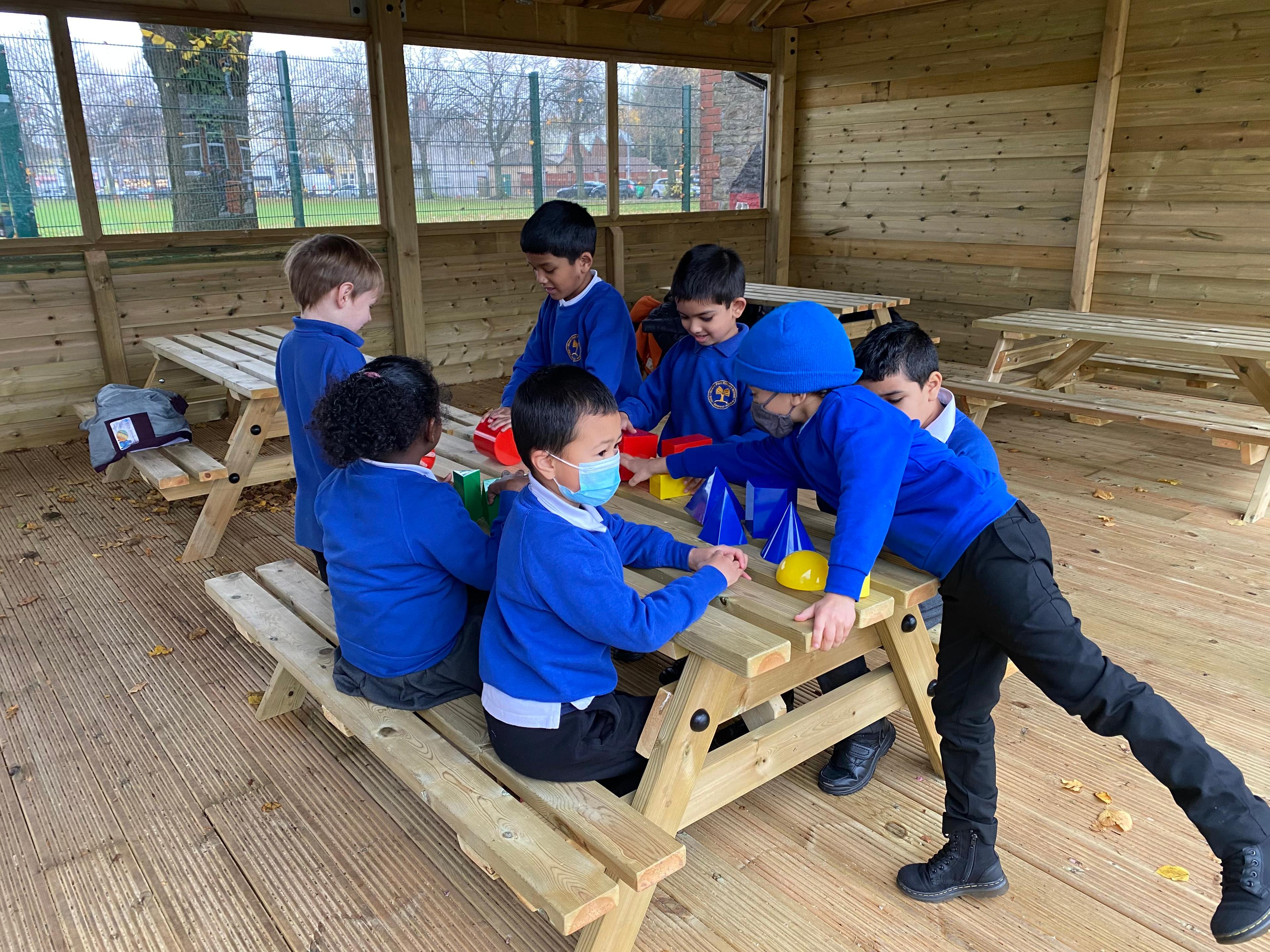 5 children sat on the picnic table, there is one young boy stood up on the side.