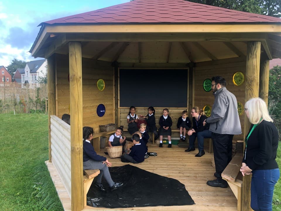 a view into the hexagonal gazebo being used as an outdoor classroom as a teacher explains a lesson to them whilst they sit on the perch benches 
