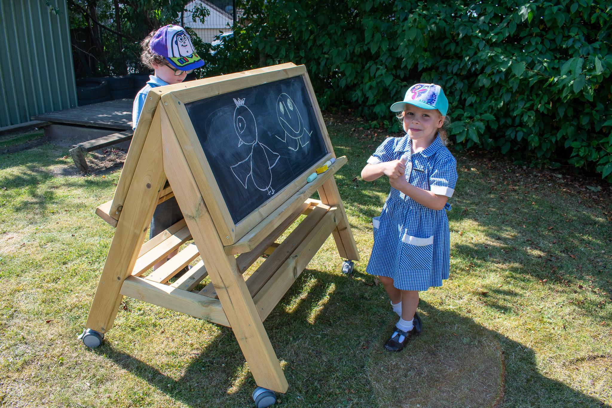 Children drawing on a wooden, chalkboard easel on wheels.