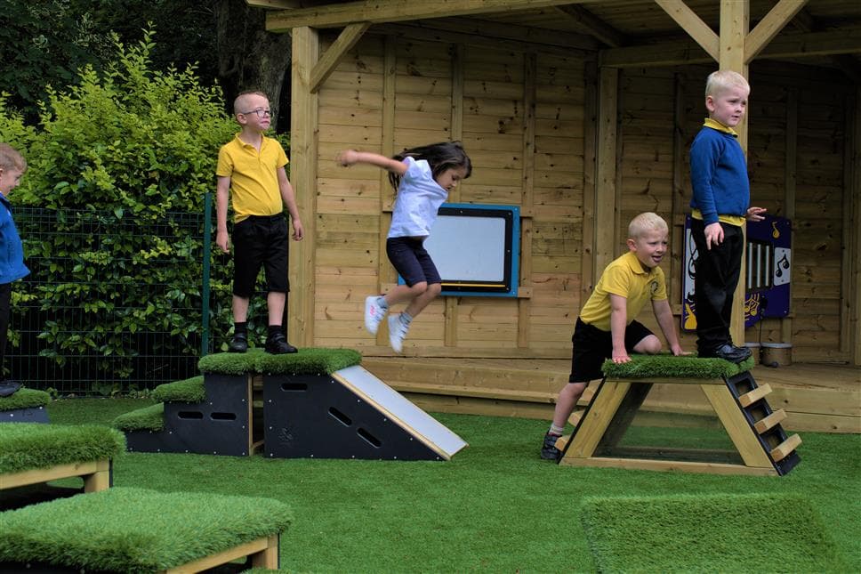 A young girl jumping off the slide in her playground, her class mates are walking around the movable blocks in her playground  