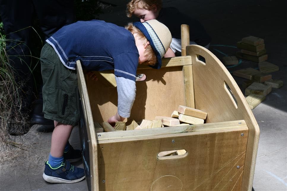 A young boy reaching into the play skip to grab the blocks inside 