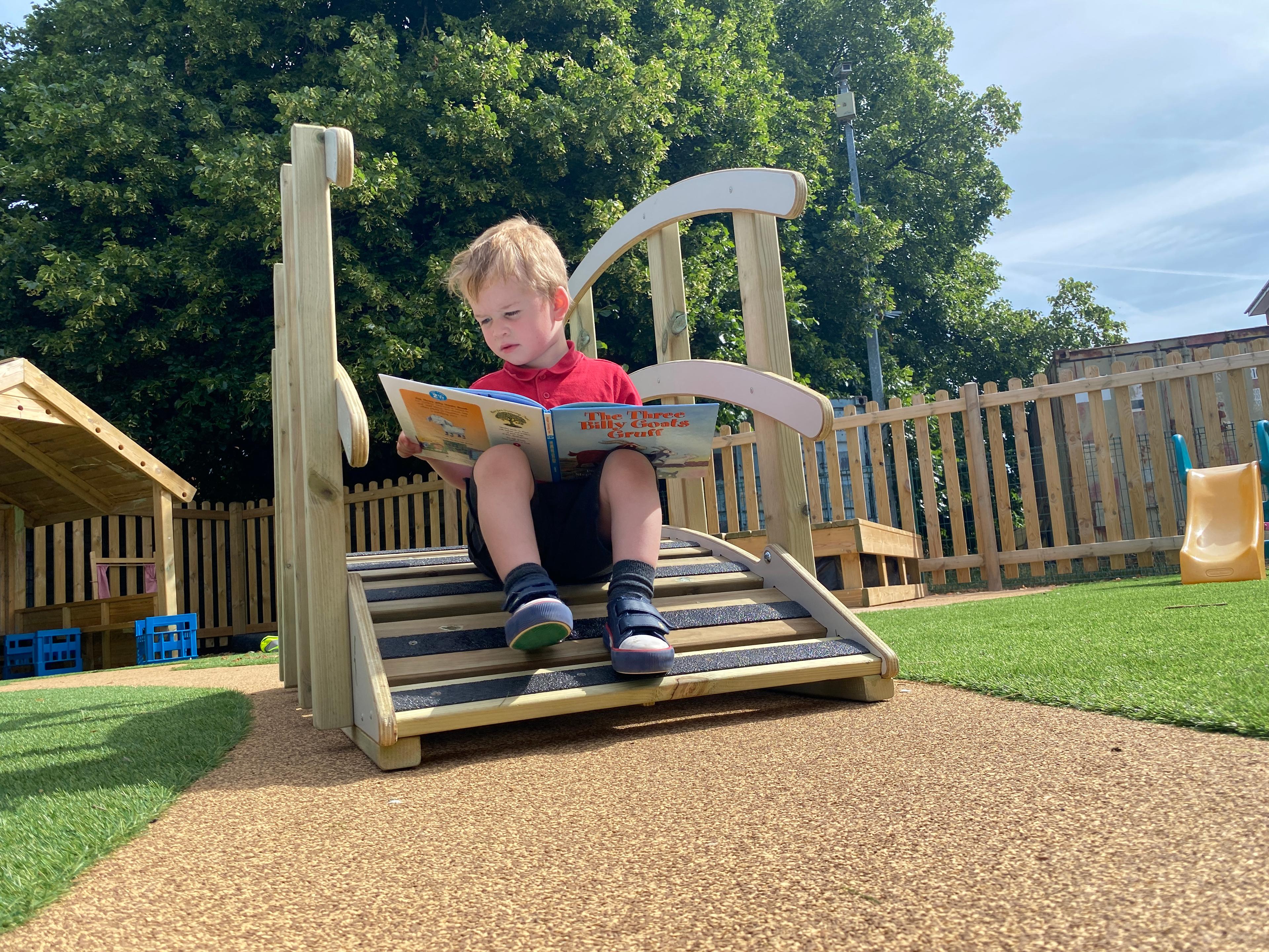 A EYFS boy sat on the end of the play bridge reading the book of the three billy goats gruff.