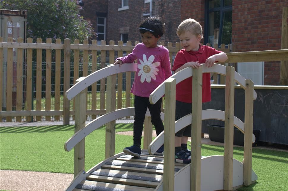 Two EYFS chldren are walking over the playbridge, they are both looking down and smiling as they walk over