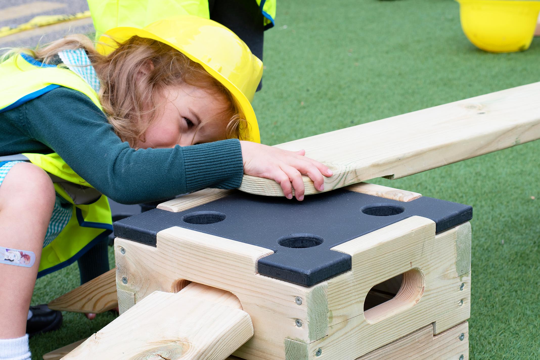 A close up of a child attaching a plank to one of the blocks from the Play Builder Technician Set. The little girl is wearing a hard hat and a high-vis jacket as she watches the parts connect.