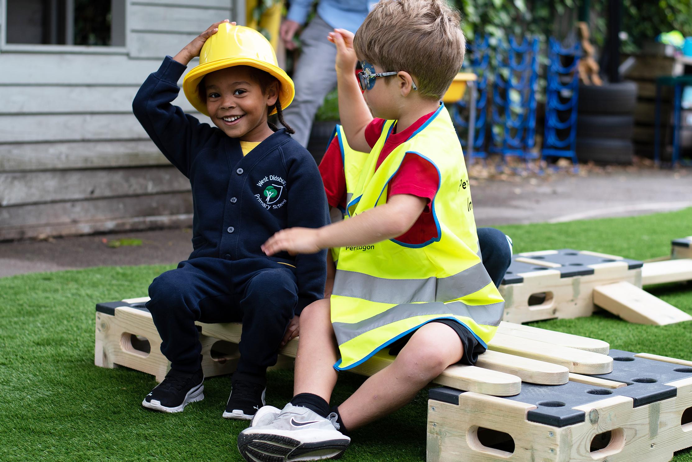 A picture of two children sat on a wooden plank from the Play Builder Technician Set. The child on the left is wearing a hard hat and the child on the right is wearing a high-vis jacket.