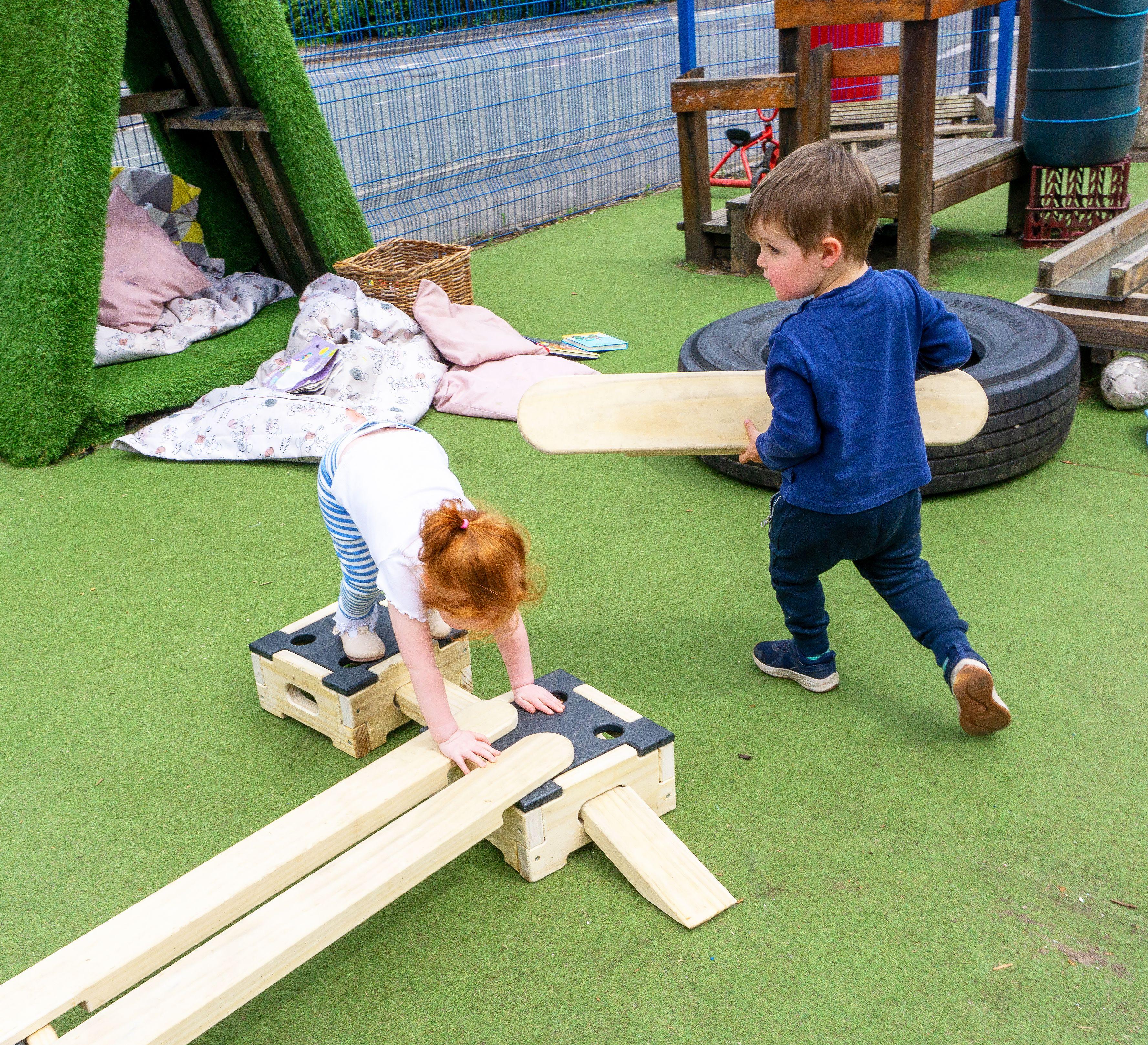 A little boy is holding a wooden plank and moving it around a little girl, who is bent over trying to bridge the gap between two wooden blocks from the Play Builder Technician Set.