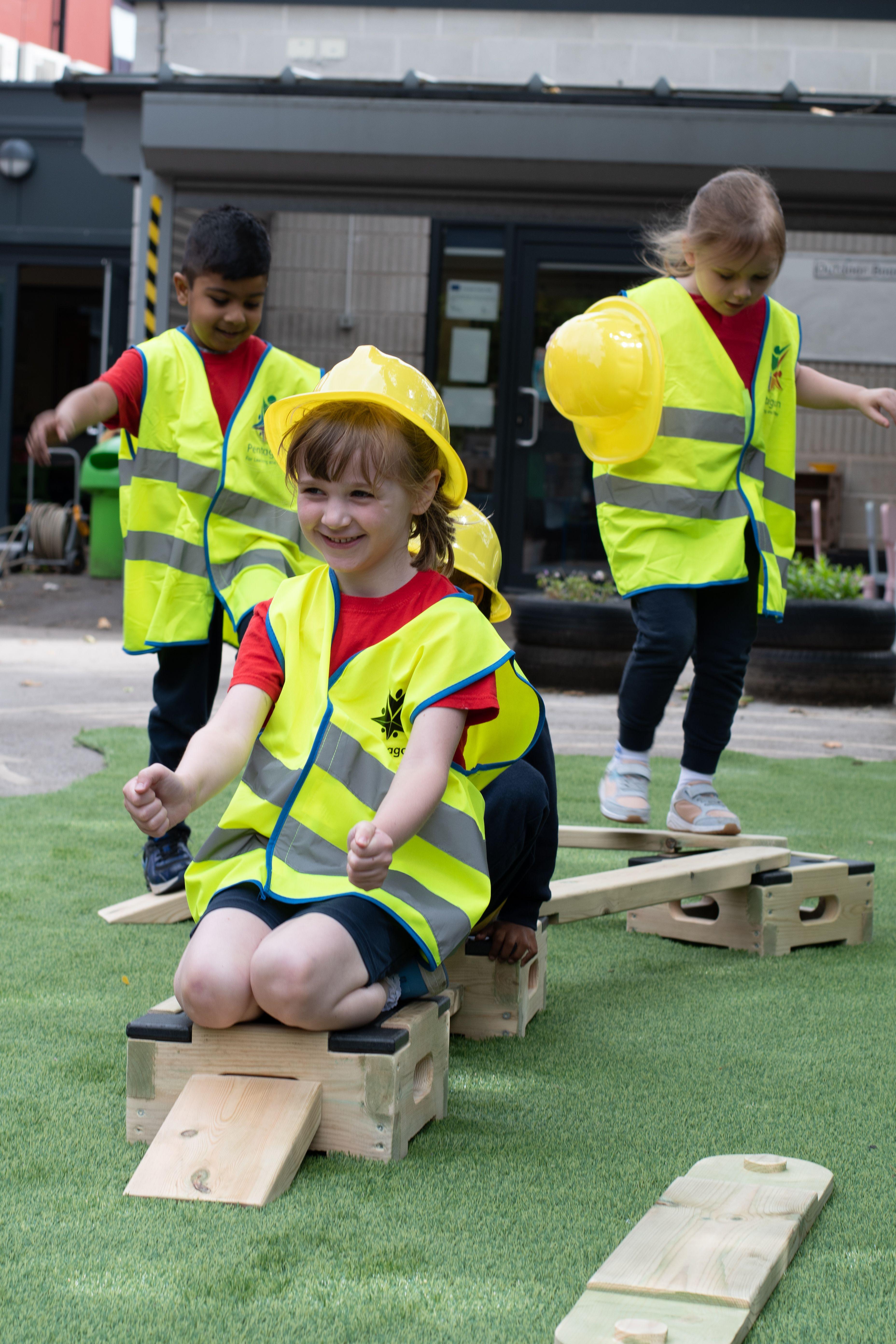 4 children are wearing hi-vis vests and builder's hats as they complete an obstacle course created from the Play Builder Technician Set. All the children are smiling as the balance across the planks.