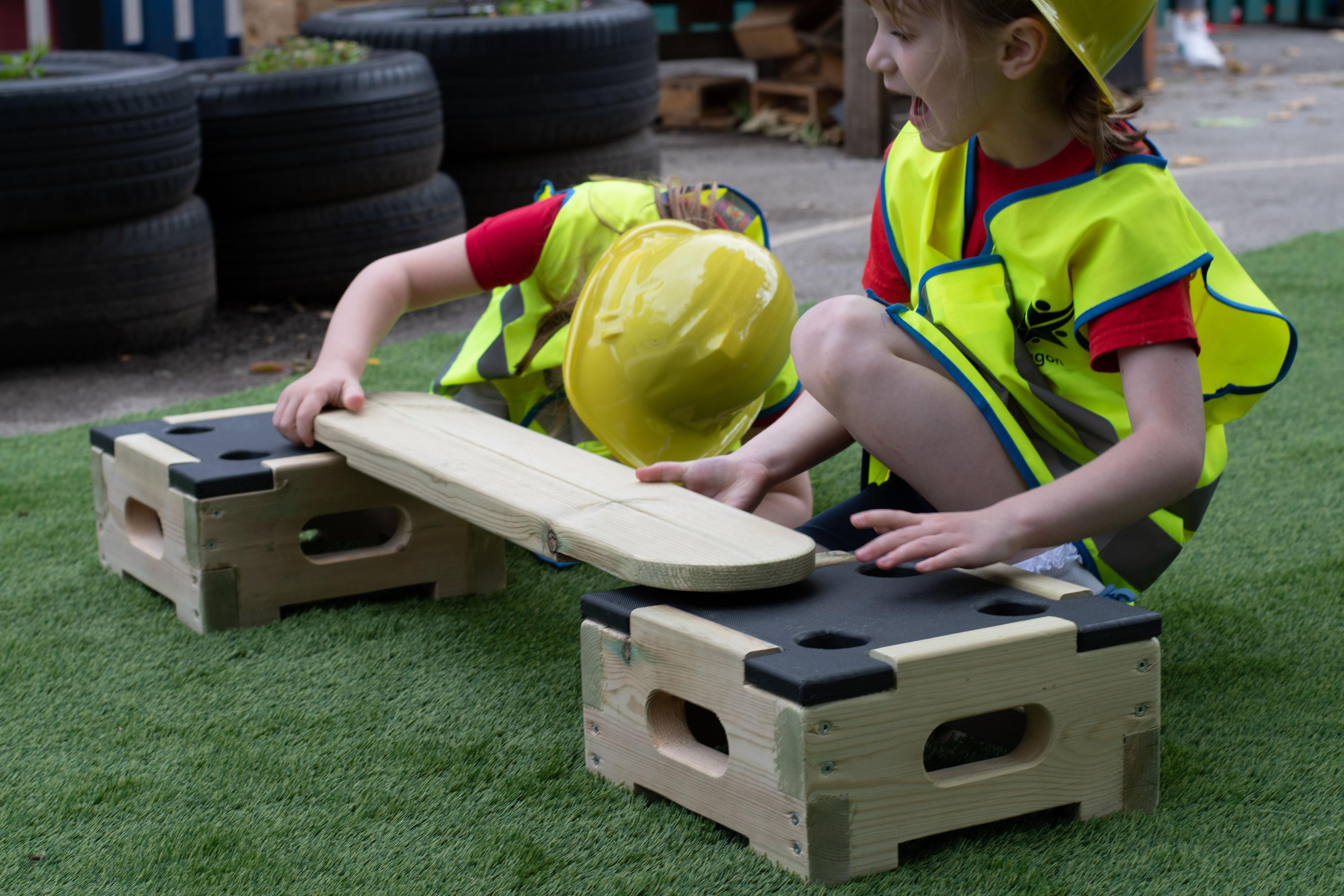 2 children wearing hi-vis vests and builder's hats are playing with the Play Builder Technician set. Both children are knelt down and placing a wooden plank between two wooden blocks, connecting them.