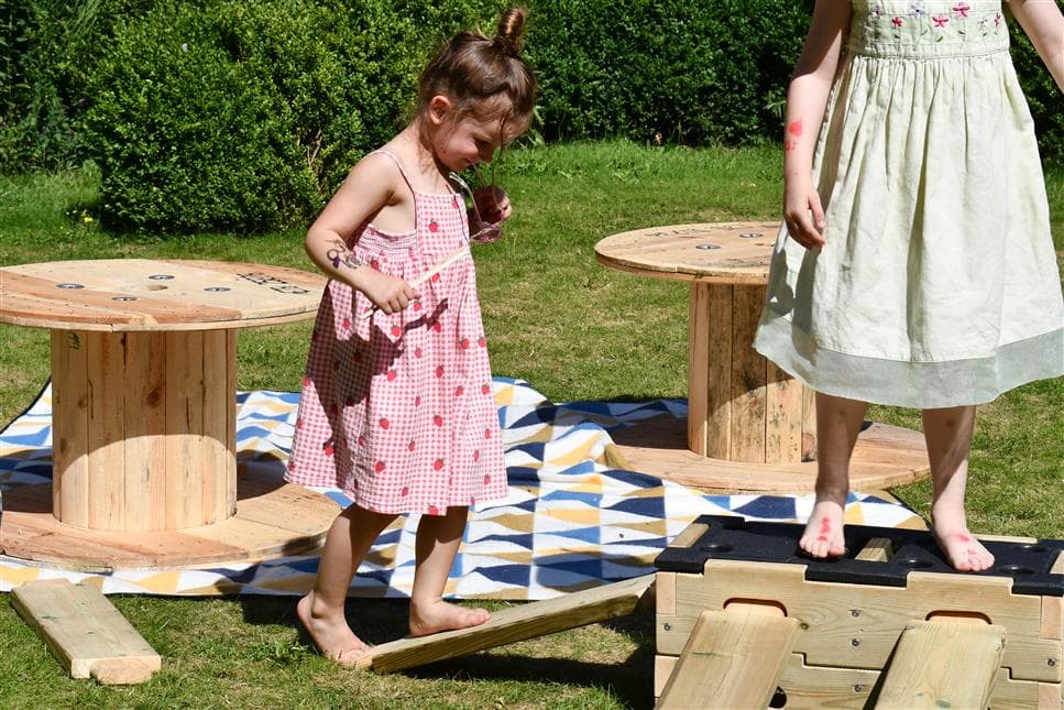 1 young girl climbing up the plank of wood and one young girl is stood on top of the play builder 