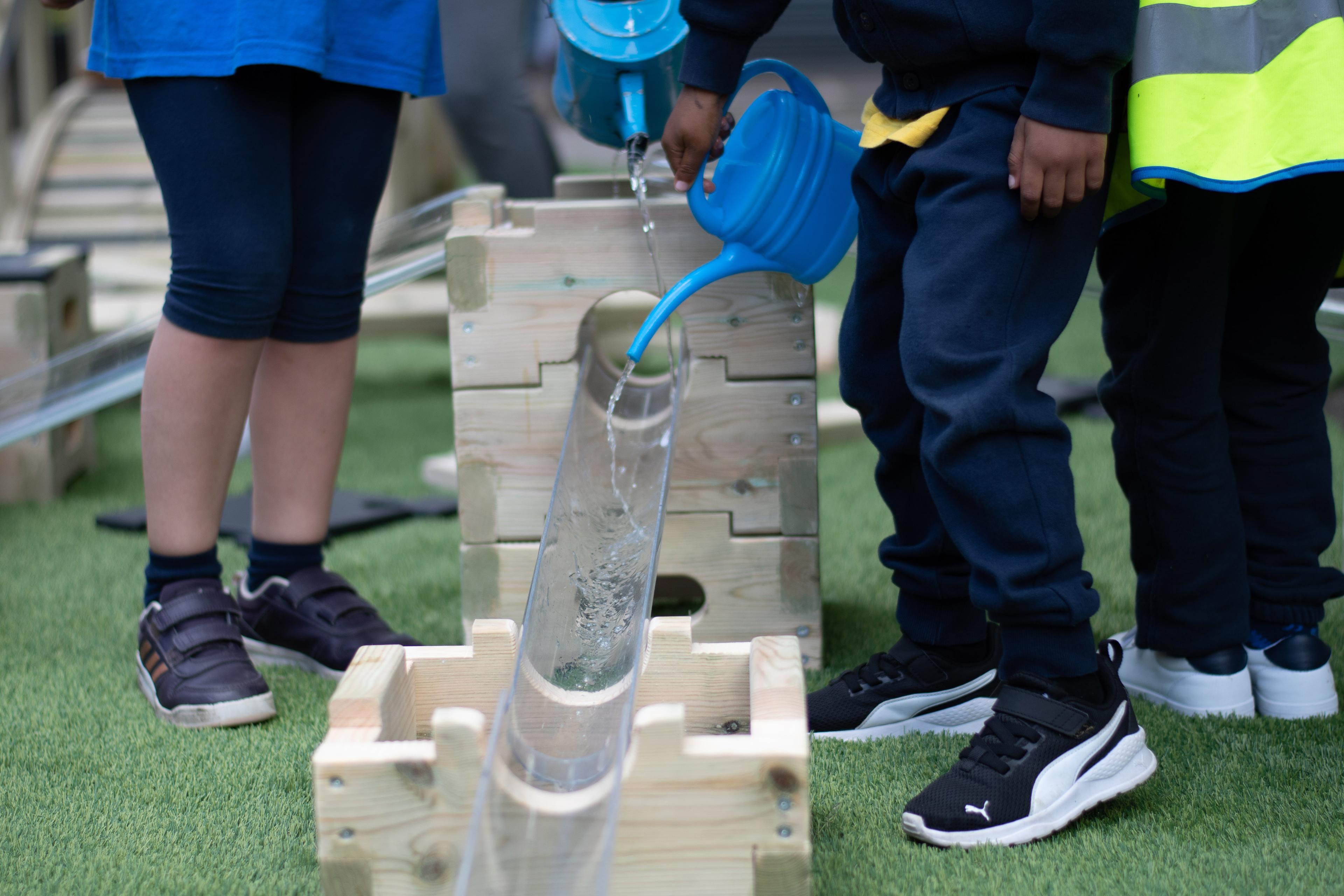 A close up of a child and a teacher pouring watering buckets into the clear plastic tubes that come with the Play Builder package.