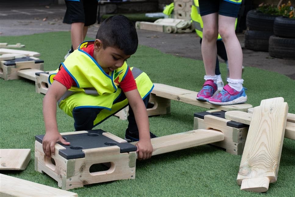 A young boy playing with the play builder set in their playground, they are wearing high vis vests
