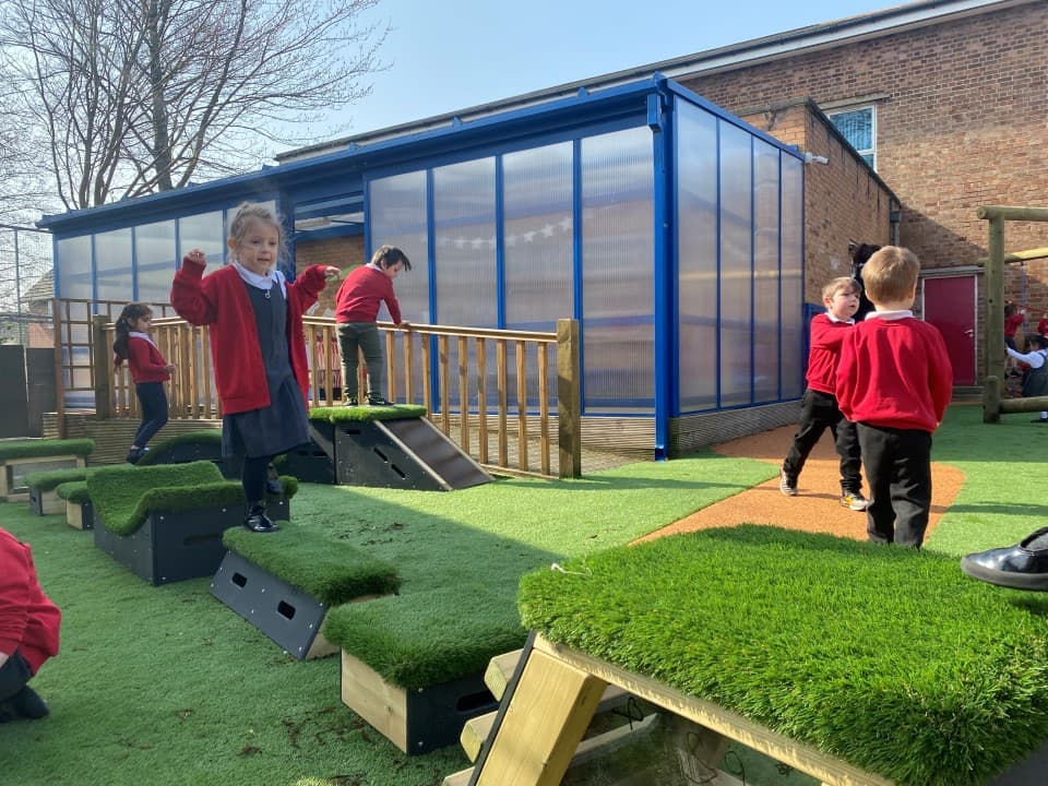 A class of children walking across the blocks in the playground, one child is leaping across