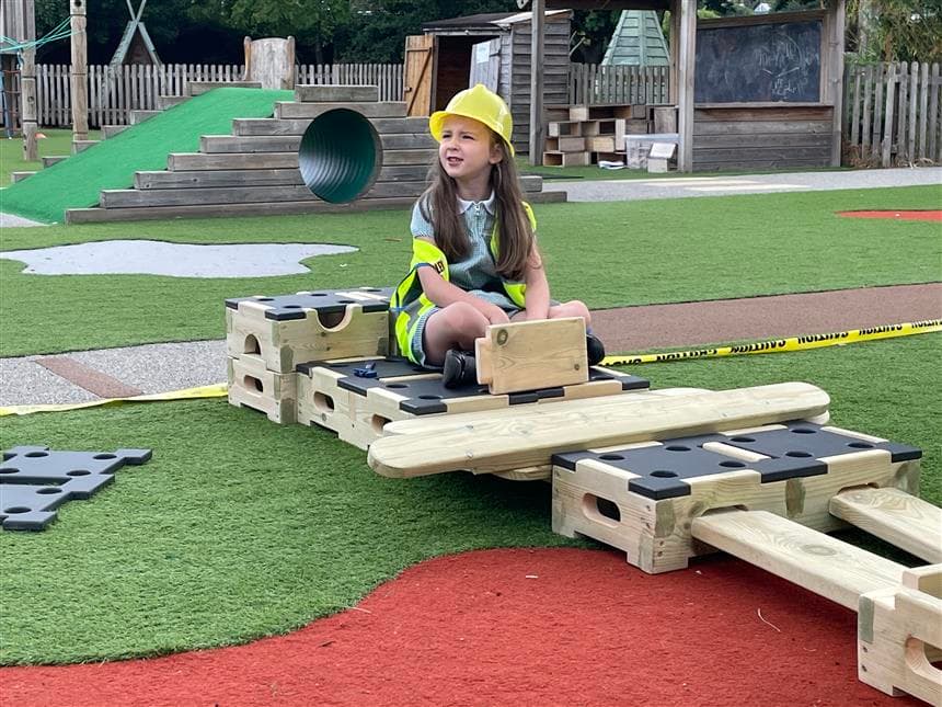 A young girl sat with a yellow hard hat on whilst using a block as a steering wheel