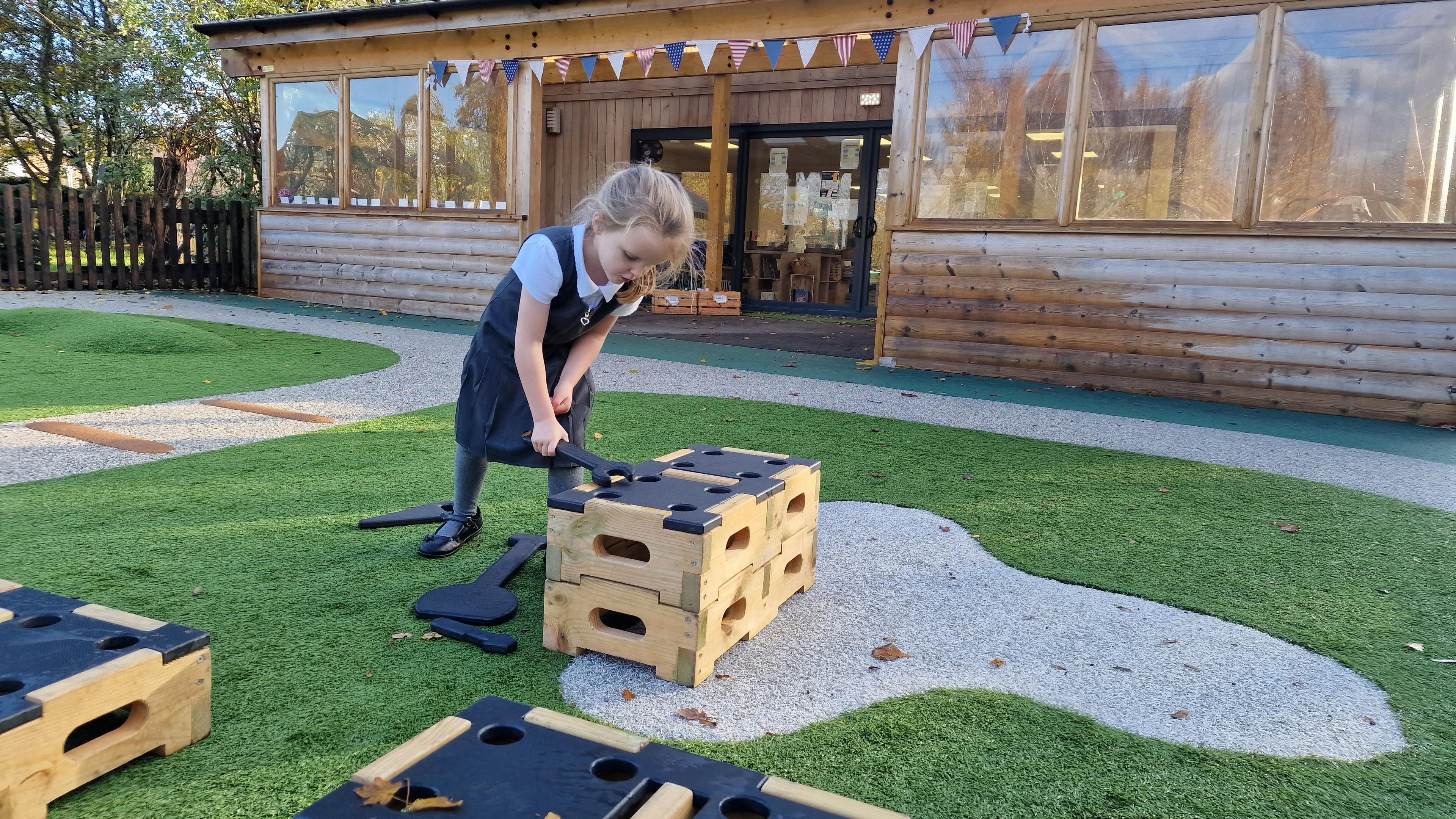 A side view of a young girl using a role play toy to saw on a block in her playground