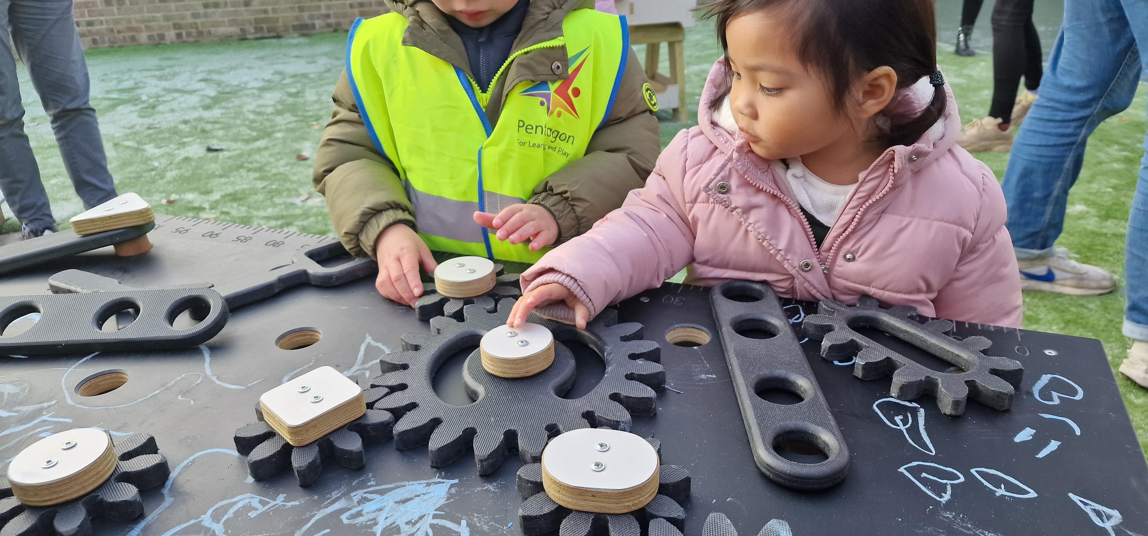 2 children playing with the gears on the construction table in their playground