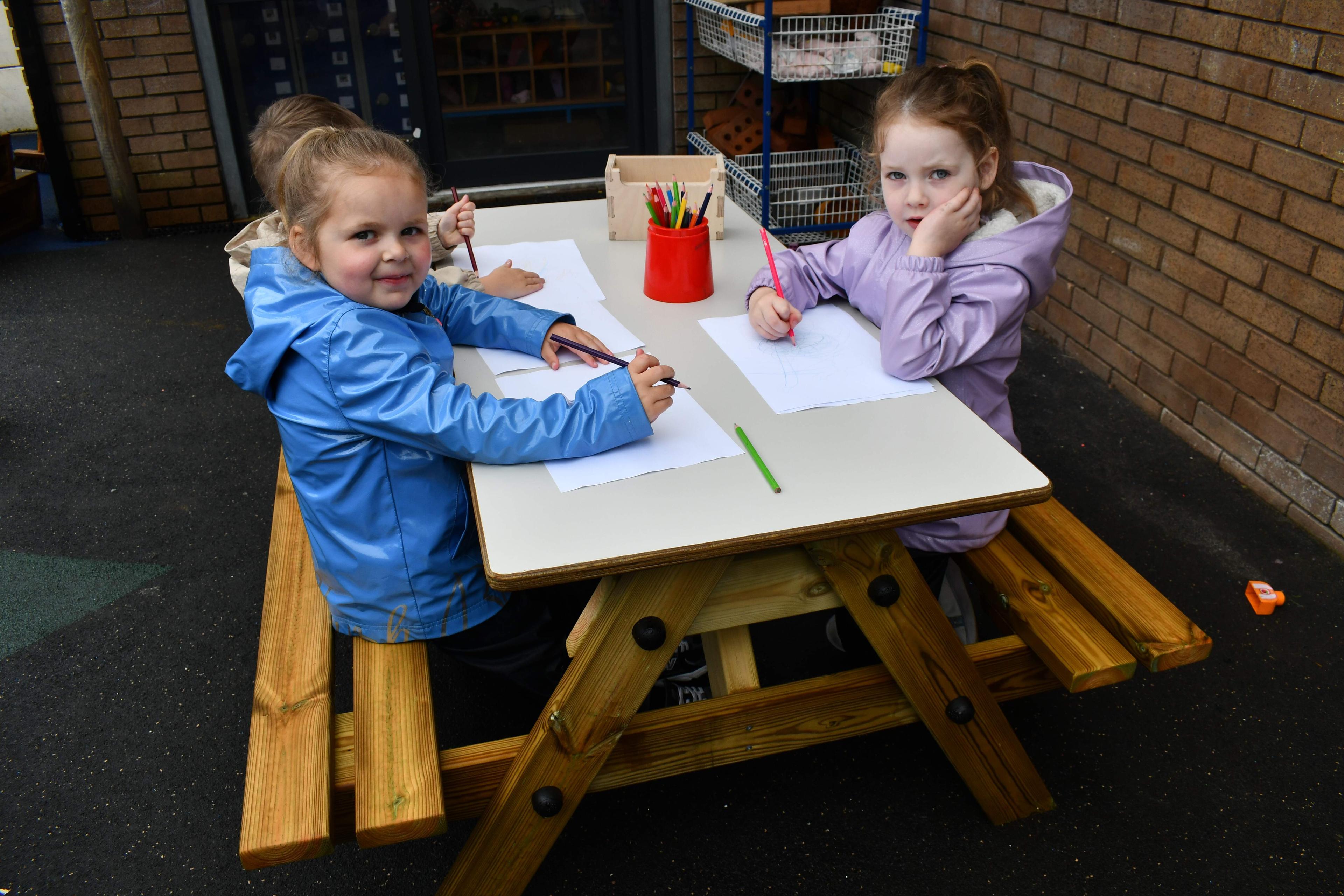 3 children sat on the Early Years Picnic Table. The children are looking at the camera, smiling, as they are drawing on paper which is placed on top of the EYFS Picnic table.