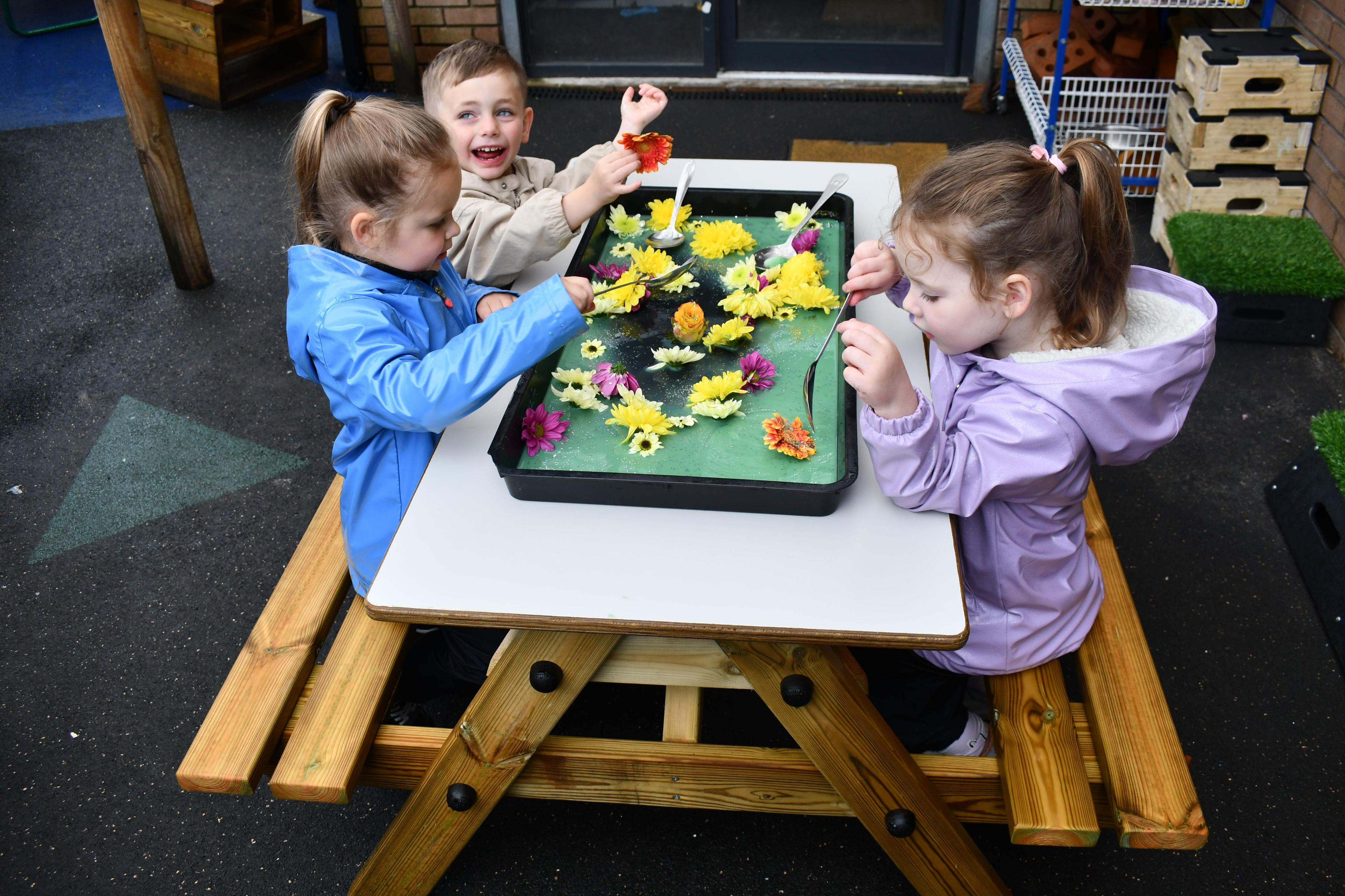 3 children are engaged with a sensory play activity that is taking place on the EYFS Picnic Table. The children are holding spoons and scooping a tray full of flowers and goo.