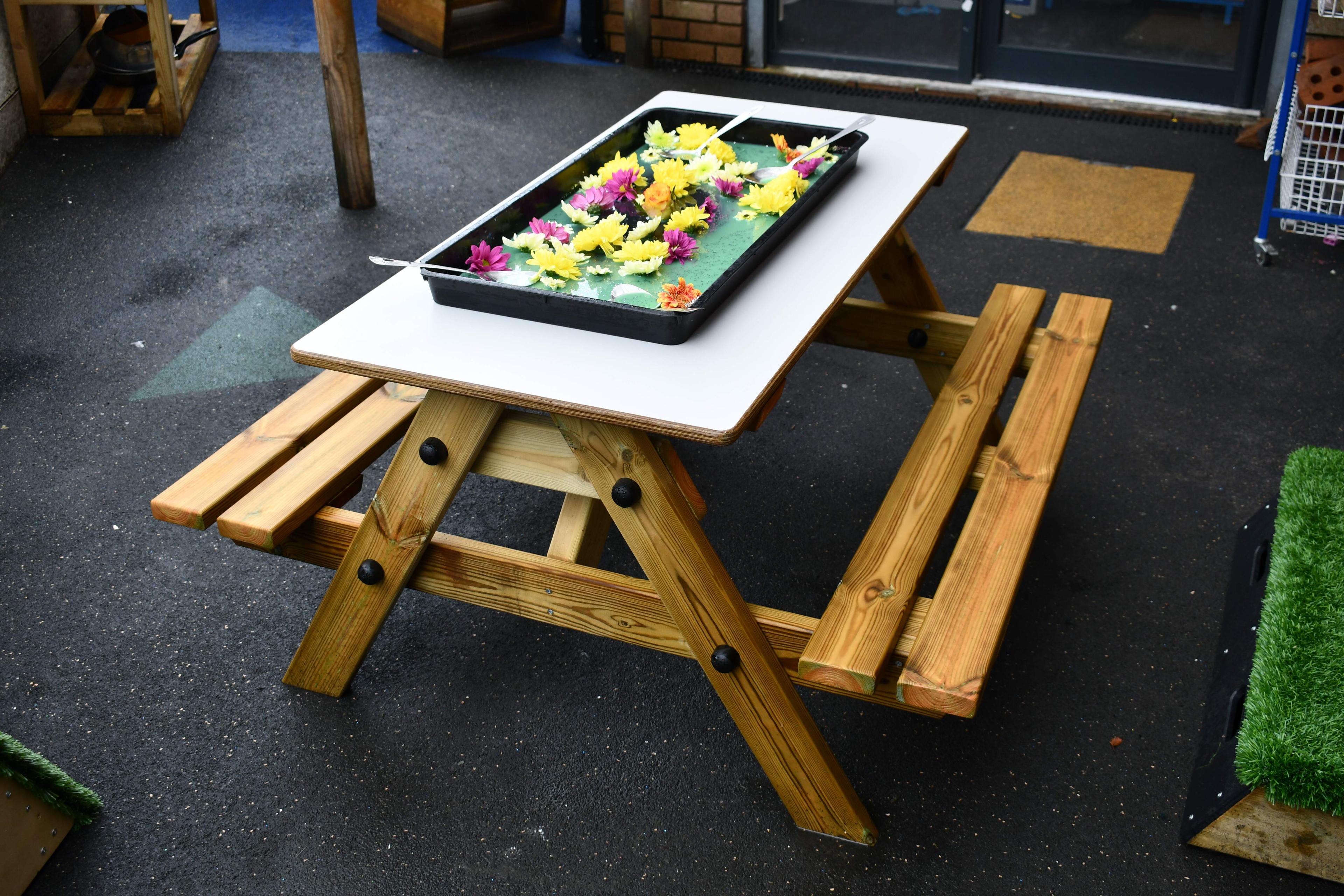 The Early Years Picnic table is set up on tarmac. A tuff tray full of flowers, green goo and spoons are placed on the grey top of the EYFS Picnic table, showing a wide variety of flower colours.
